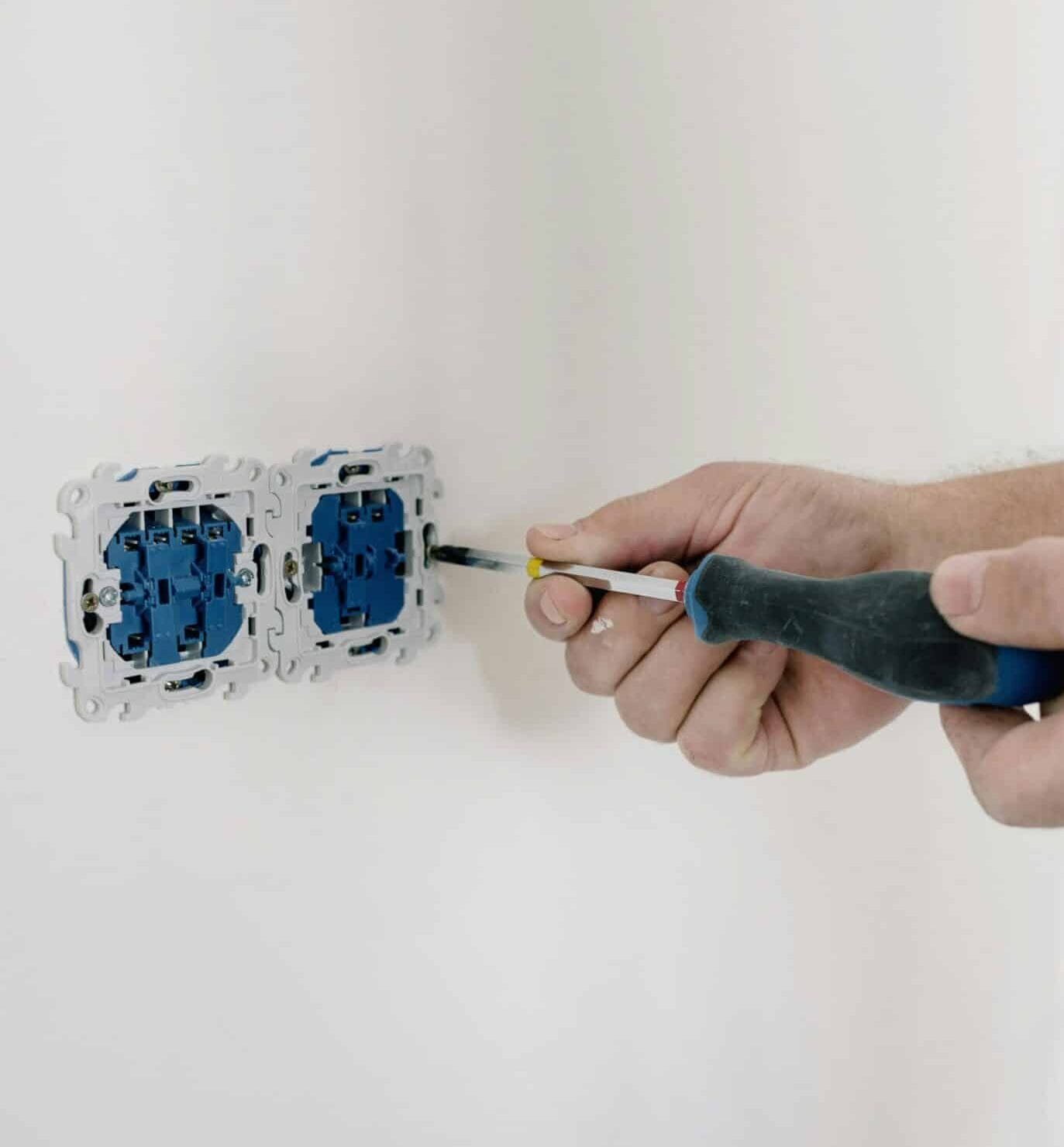 Close-up van de handen van een professionele elektricien in Assen die een dubbele wandschakelaar installeert met een schroevendraaier op een witte muur.